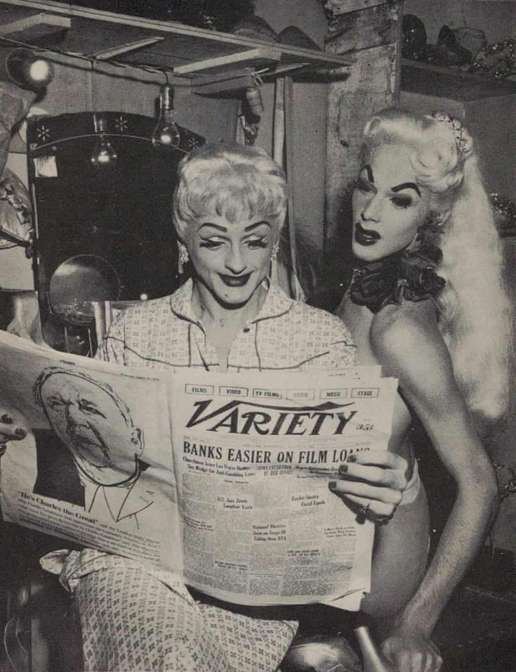 Two people reading the news paper. One person is peaking over the shoulder of the other to read he article. Their faces look amused and shocked.